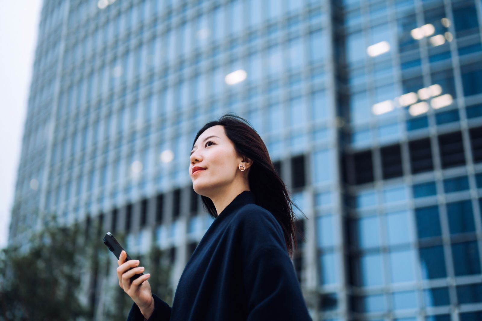 Confident and determined young Asian businesswoman looking ahead with smile, holding smartphone while commuting in downtown district against contemporary corporate skyscrapers in city. Business lifestyle with technology, staying connected anytime anywhere