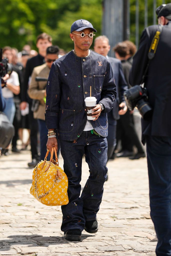 PARIS, FRANCE - JUNE 23: Pharrell Williams wears a navy blue denim with large white embroidered pattern cap, silver and gold rhinestones small circle sunglasses from Louis Vuitton, gold chain necklaces, a navy blue denim with multicolored embroidered small pattern buttoned shirt from Louis Vuitton, matching navy blue denim with multicolored embroidered small pattern large pants from Louis Vuitton, gold and diamonds bracelets, gold rings, a white t-shirt, black suede and leather sneakers from Louis Vuitton,